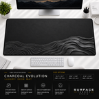 Charcoal Evolution desk mat beneath a monitor on a bright desk with a white keyboard and mouse.