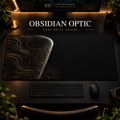 Obsidian Optic desk mat, near-black with bronze-gold contours in a rounded form on the left, in a warm, dark workspace.