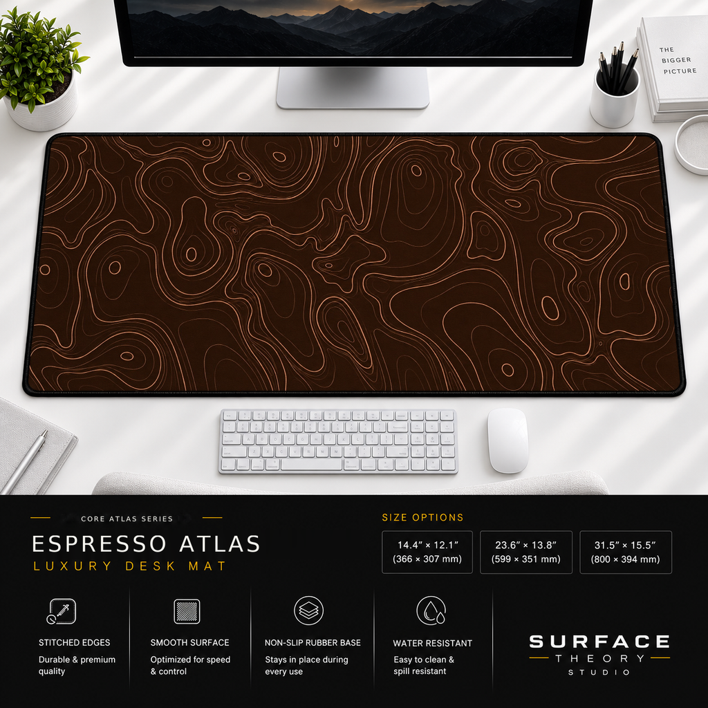 Espresso Atlas desk mat beneath a monitor on a bright desk with a white keyboard and mouse.