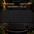 Graphite Atlas desk mat with fine topographic-inspired contours across the full surface in a warm, dark workspace.